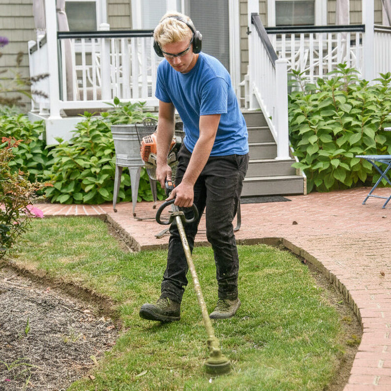 Professional worker using a string trimmer to create clean edges along a residential brick walkway.
